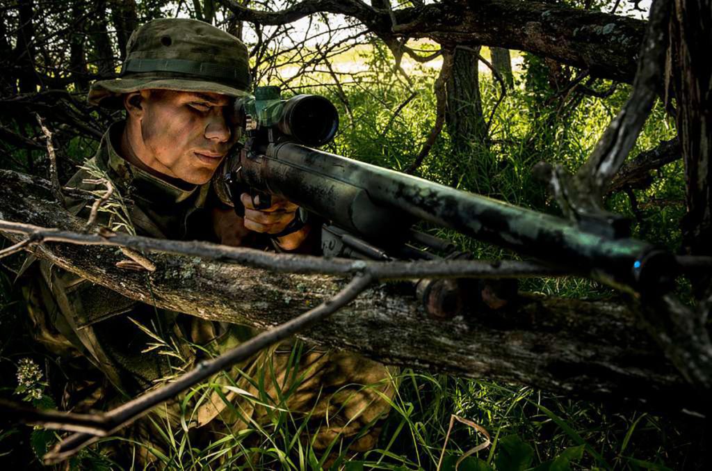 a sniper wearing a jungle hat looks through a rifle scope